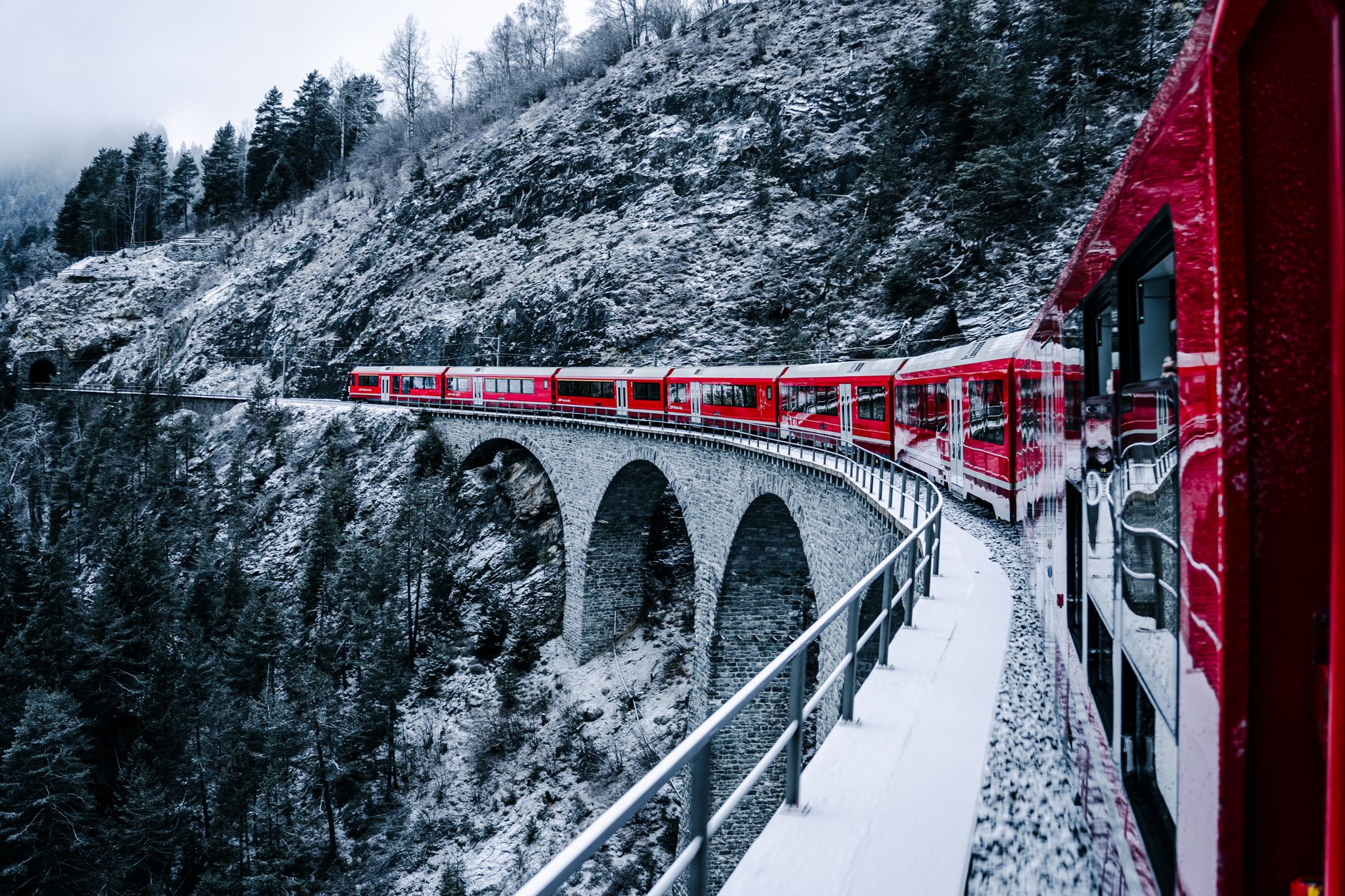 Červený panoramatický vlak Glacier Express projíždí přes kamenný viadukt v zasněžených švýcarských horách